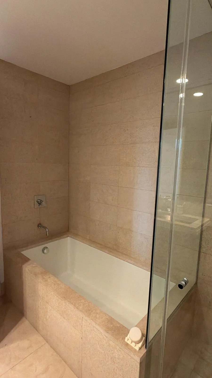 Deep white soaking tub framed in beige stone tile with a wall-mounted chrome spout and glass shower wall beside it.
