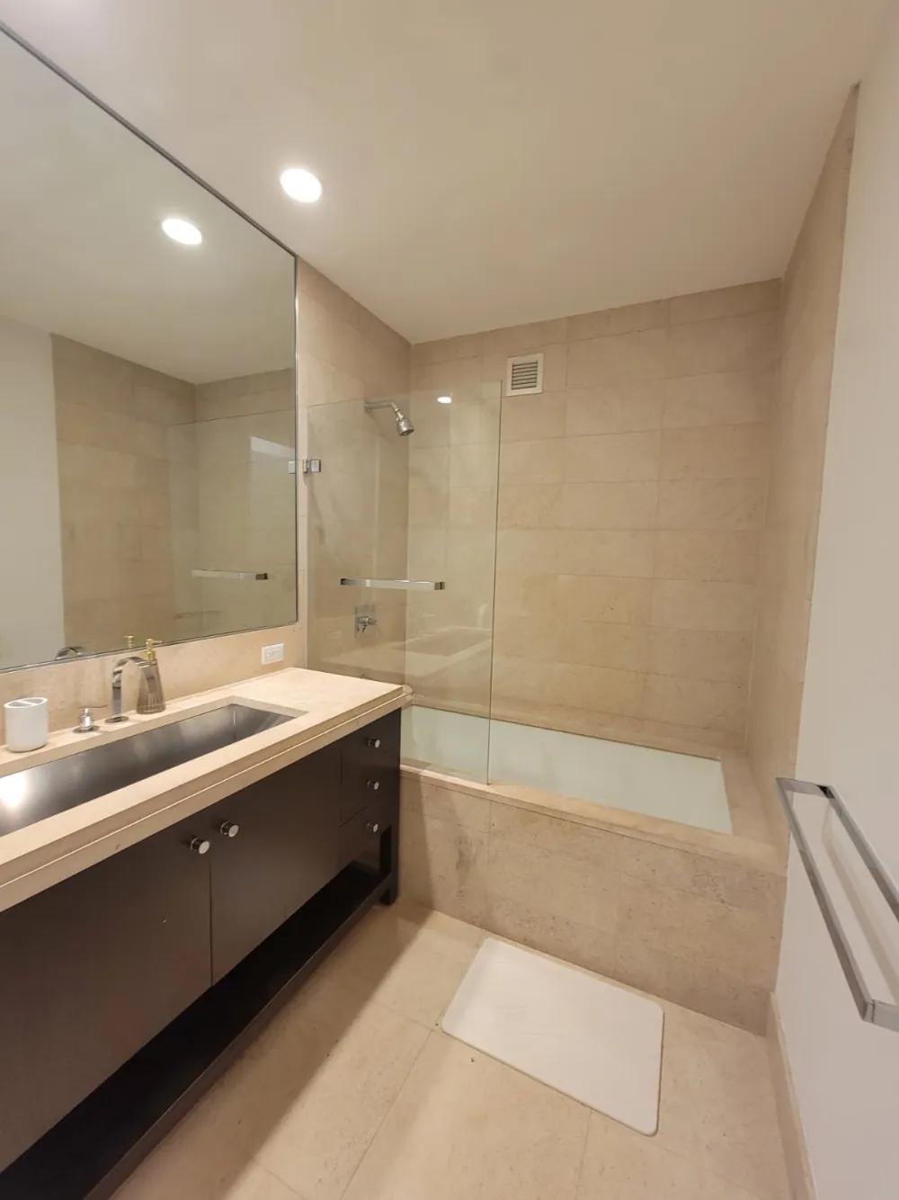 Bathroom with beige stone tile, tub with glass panel, dark wood vanity with steel trough sink and wall mirror.