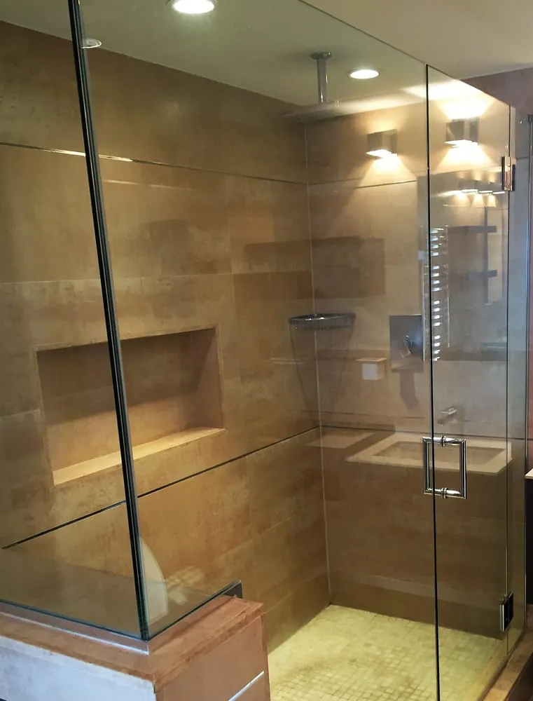 Frameless glass shower with large tan porcelain tile, long niche, ceiling rain head and beige mosaic floor.