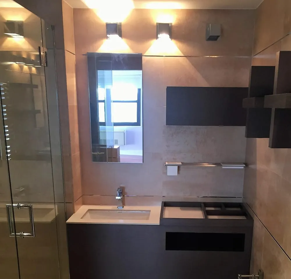 Bathroom with tan porcelain tile, dark wood floating vanity and shelves, white sink top and cube sconces.