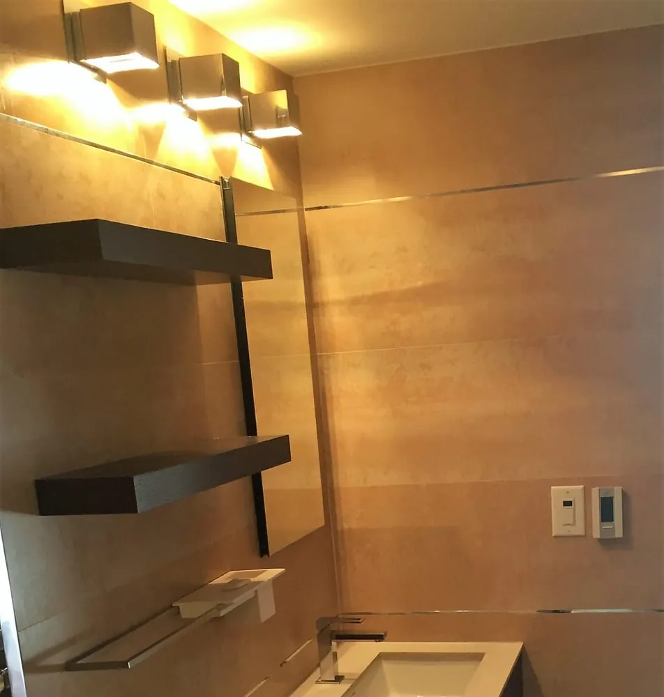 Bathroom wall with tan porcelain tile, dark wood floating shelves, cube sconces and a white vanity top.