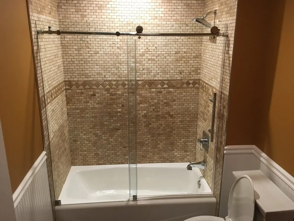 Bathtub with tan travertine mosaic surround, diamond border, sliding glass door, white beadboard wainscot and orange walls.