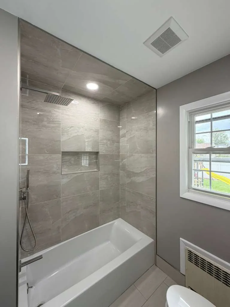 Alcove tub with gray marble-look porcelain tile to the ceiling, a niche, rain shower head and glass panel.