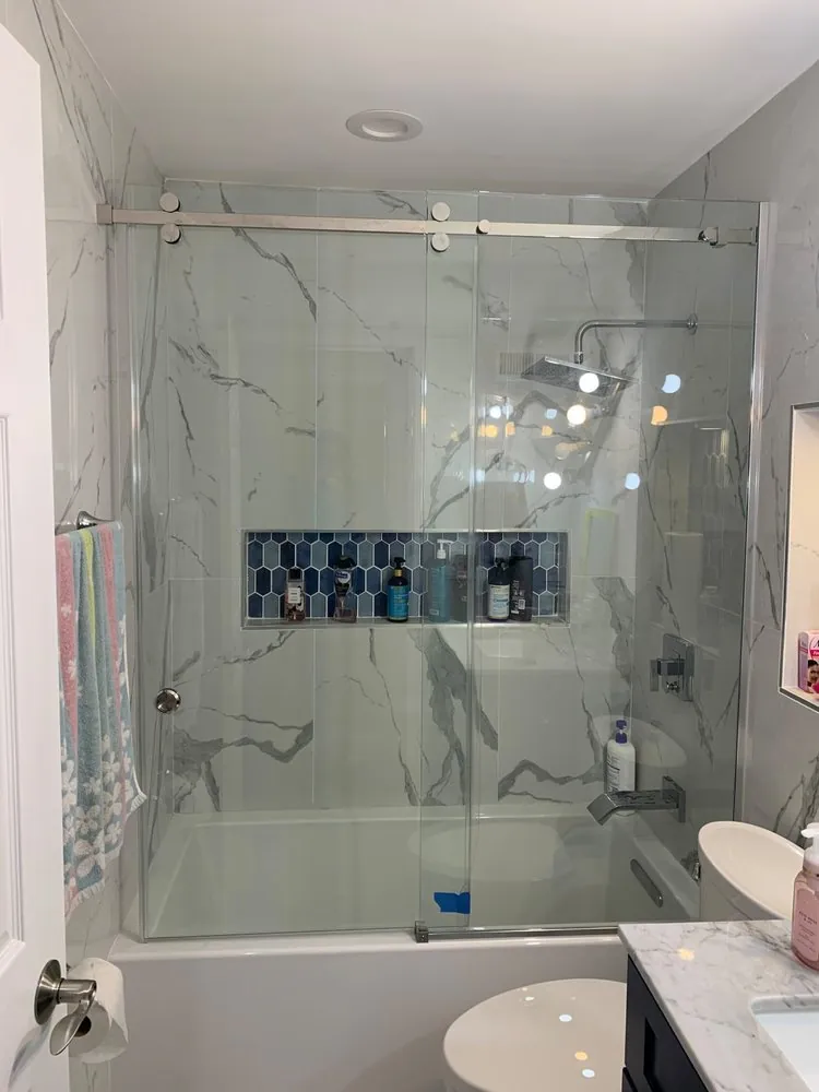 Tub with frameless sliding glass door, white marble-look porcelain walls, blue hex mosaic niche and a navy vanity.