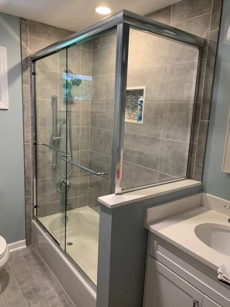 Bathtub with sliding glass doors, gray large-format wall tile, mosaic niche, white vanity and gray plank floor.