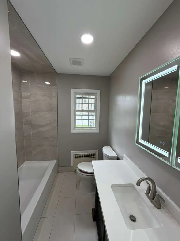 Narrow bathroom with tiled tub alcove on the left, white vanity top and lighted mirror on the right, toilet under window.