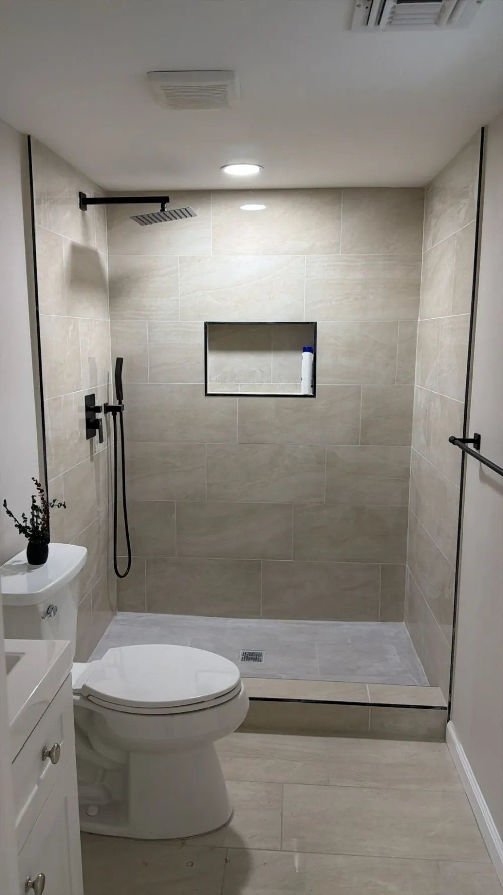 Walk-in shower with large beige porcelain tile, black rain showerhead and handheld, recessed niche, white toilet and vanity.