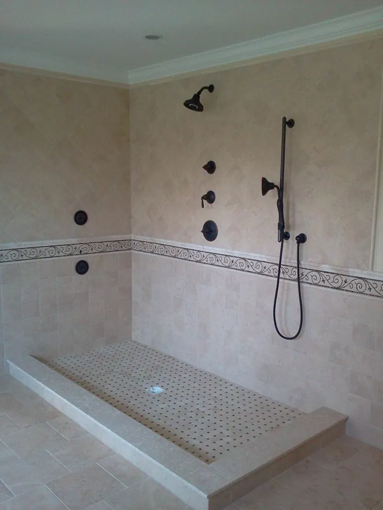 Large open walk-in shower with beige tile, scroll-pattern border, basketweave floor and oil-rubbed bronze fixtures.