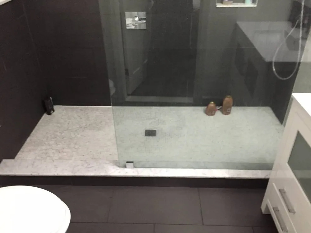 Walk-in shower with marble mosaic floor, dark wall tile and glass panel next to a white vanity.