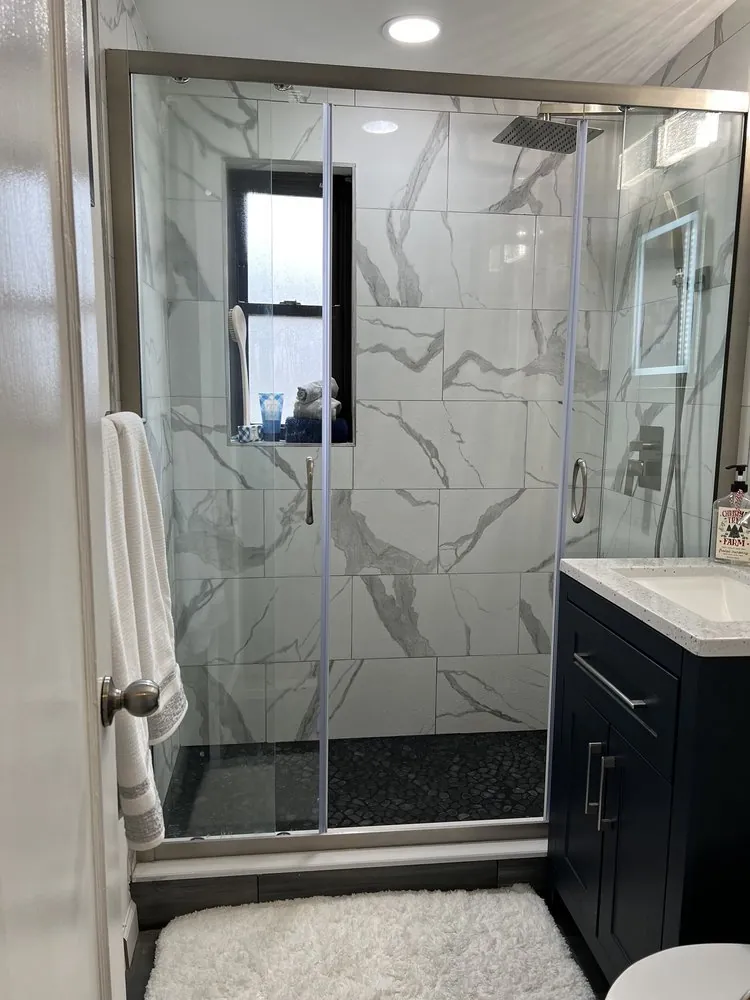 Walk-in shower with white marble-look tile, window niche, black pebble floor and brushed nickel sliding glass door.