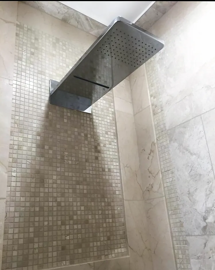 Rectangular chrome waterfall rain shower head mounted on cream marble tile with a small square mosaic strip.