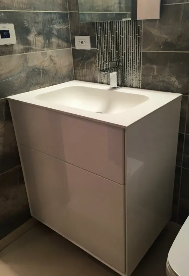 White high-gloss vanity with integrated sink and chrome faucet against dark stone-look wall tile and a glass mosaic strip.
