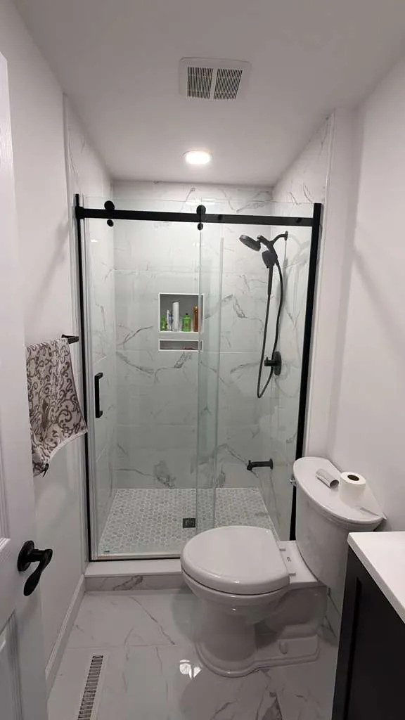 Small bathroom with white marble-look tile shower, hexagon mosaic floor, black sliding glass door and black fixtures.