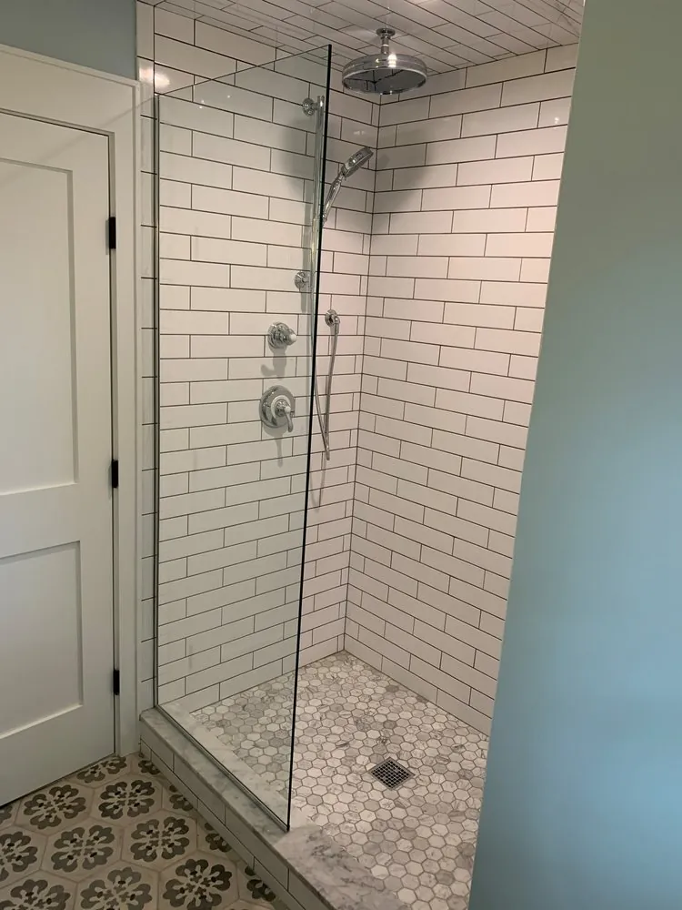 Shower with white subway tile and dark grout, glass panel, marble hexagon floor and patterned floor tile outside.