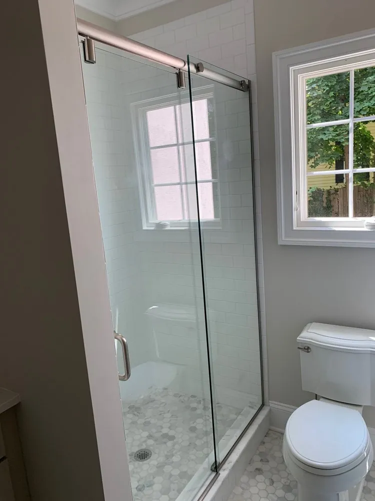 Shower with white subway tile, marble hexagon floor and a sliding glass door on a brushed rail, next to a white toilet.