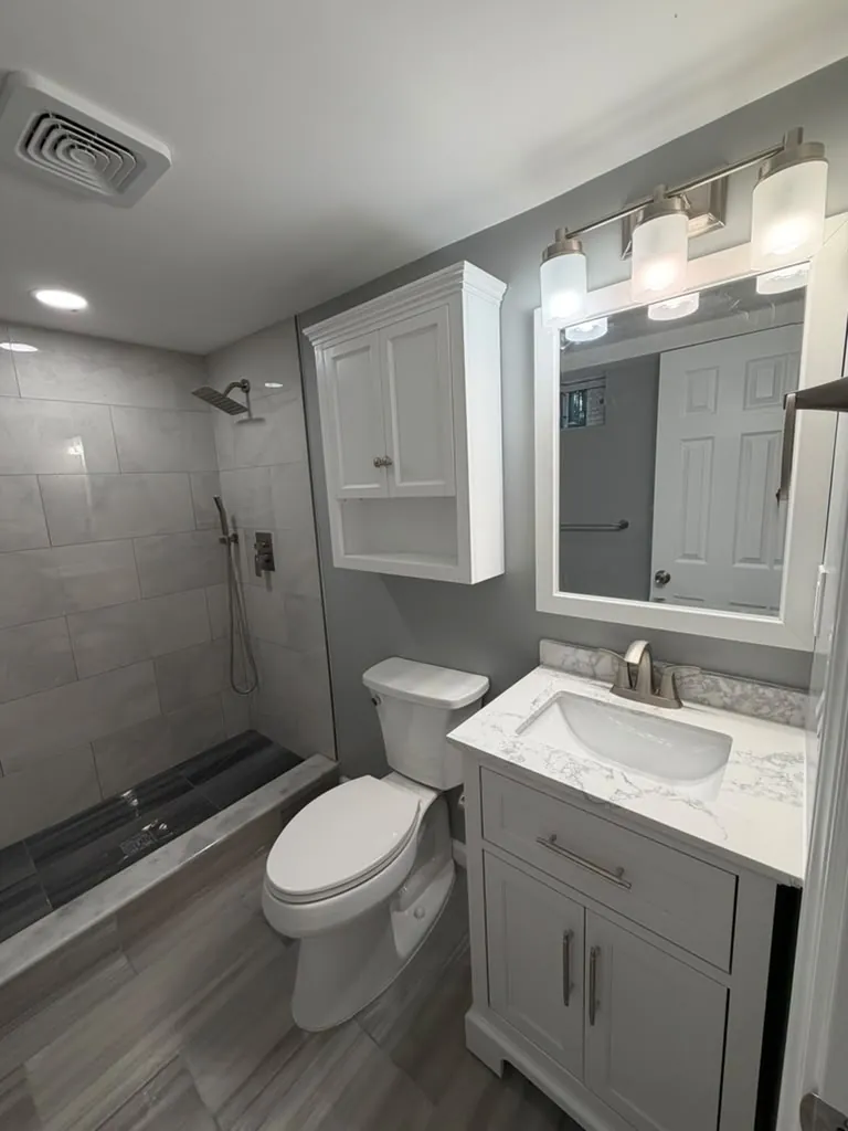 Bathroom with white vanity, marble-look top, white framed mirror, three-light fixture, over-toilet cabinet and gray walls.