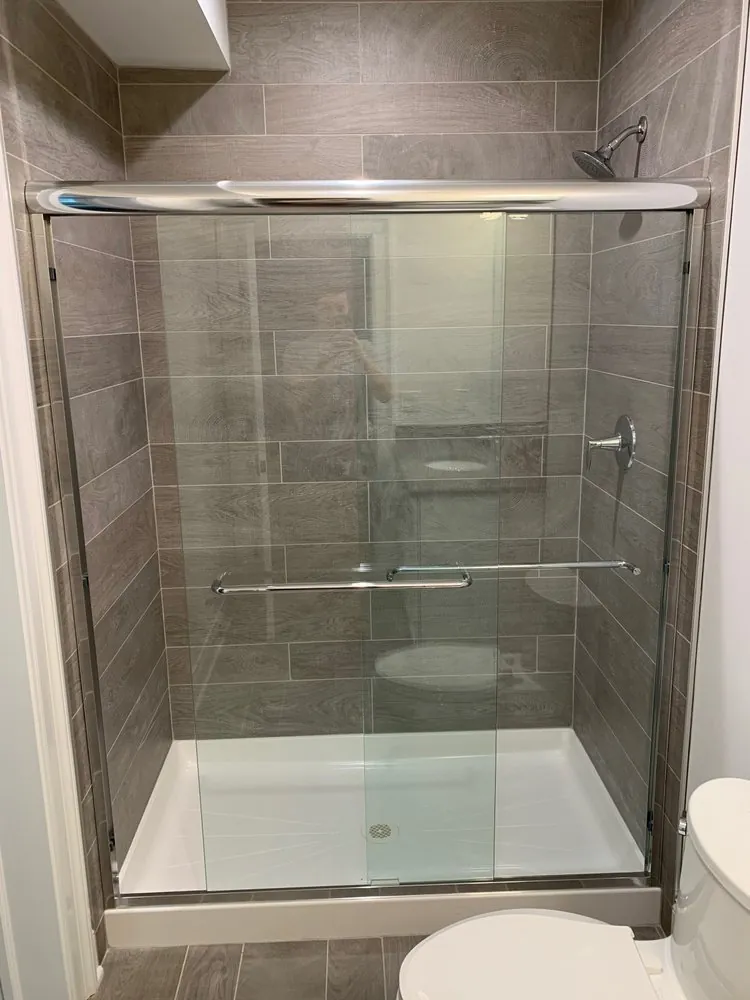 Shower with gray wood-look plank tile, white acrylic base and chrome-framed sliding glass doors with towel bars.
