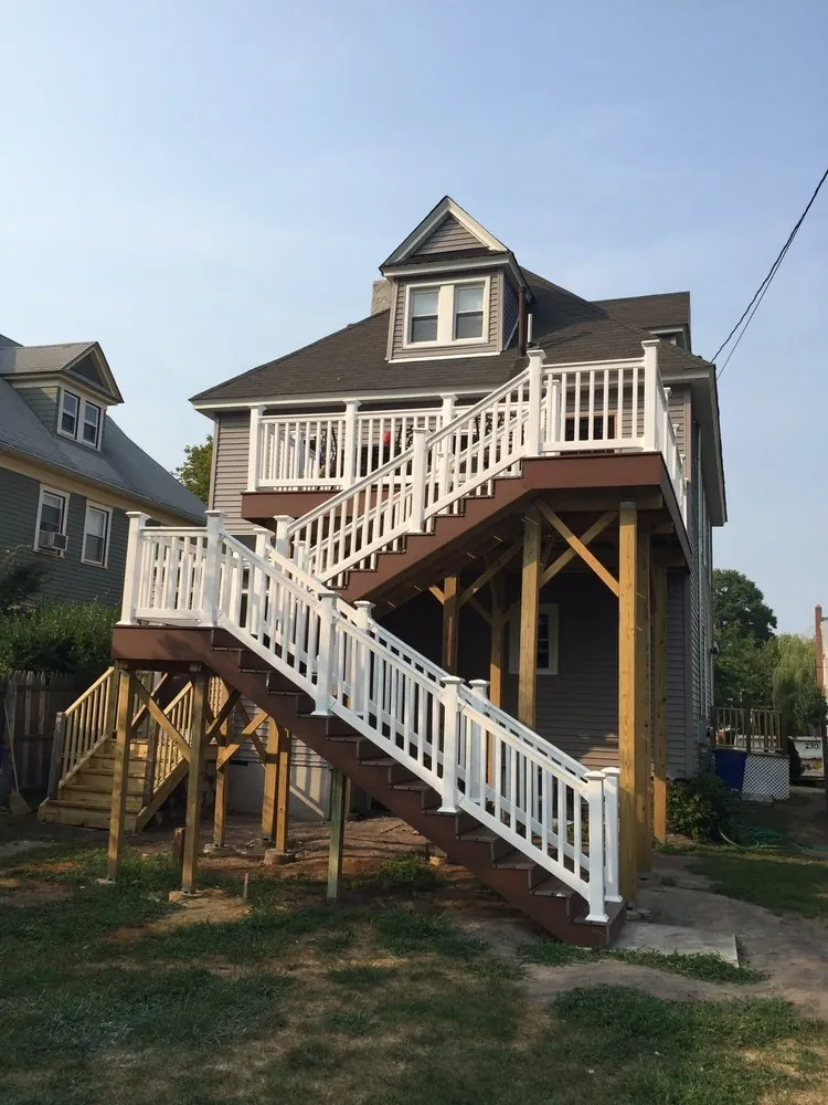 Elevated rear deck with white rails and a long brown staircase on pressure-treated posts behind a gray sided house.