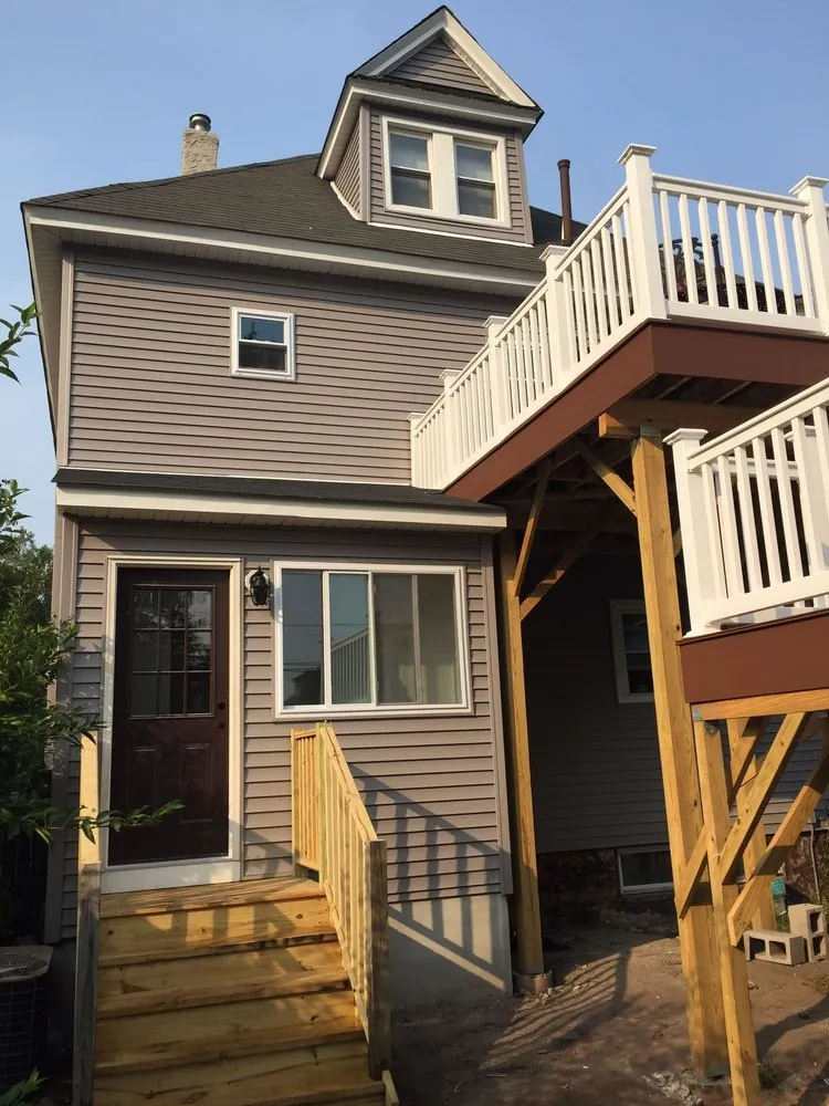 Rear of a gray house with a new wood back-door stoop and an upper deck with white railings on wood posts.