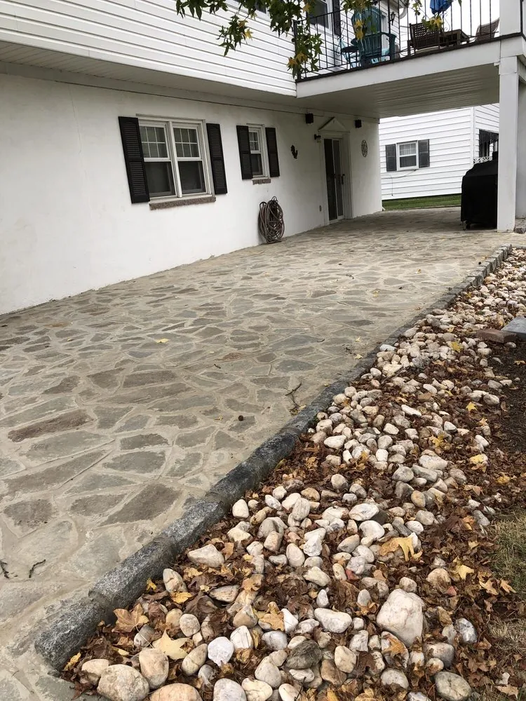 Irregular gray flagstone patio along a white stucco wall with a curb edge and a river rock border under a deck.