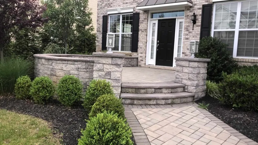 Paver front patio with curved block steps, stone seat walls and lamp pillars leading to a black front door.