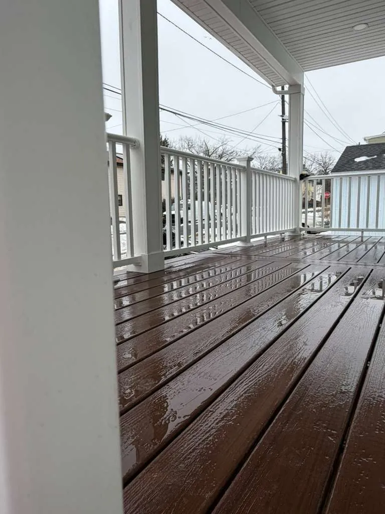 Low view across wet brown composite porch boards toward white columns and vinyl railing under a covered roof.