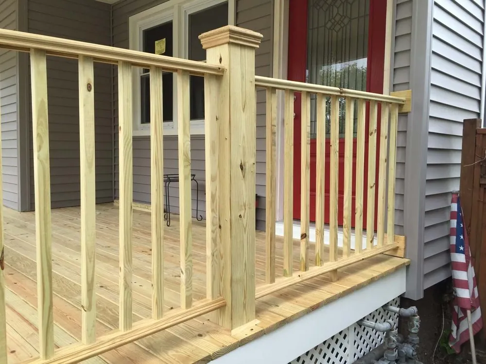 Close view of new pressure-treated wood porch railing and decking in front of a red door and gray siding.