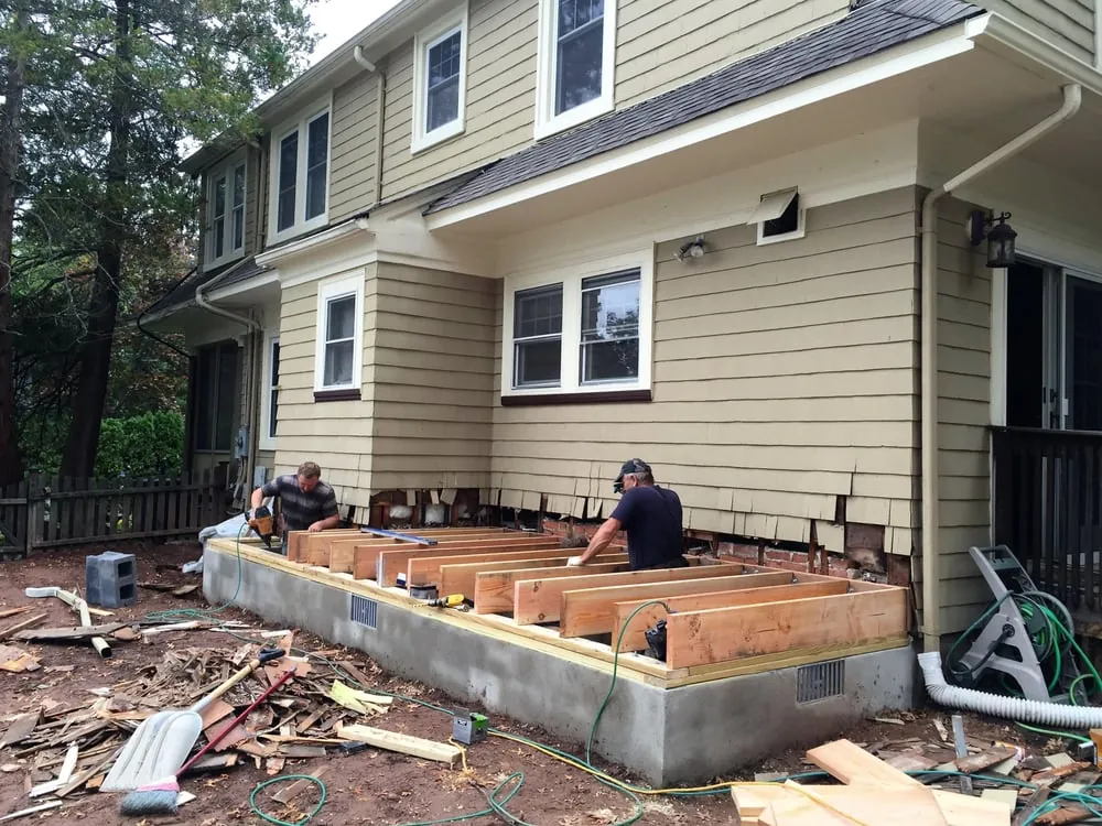 Two workers setting wood floor joists on a new concrete block foundation against a tan sided house.