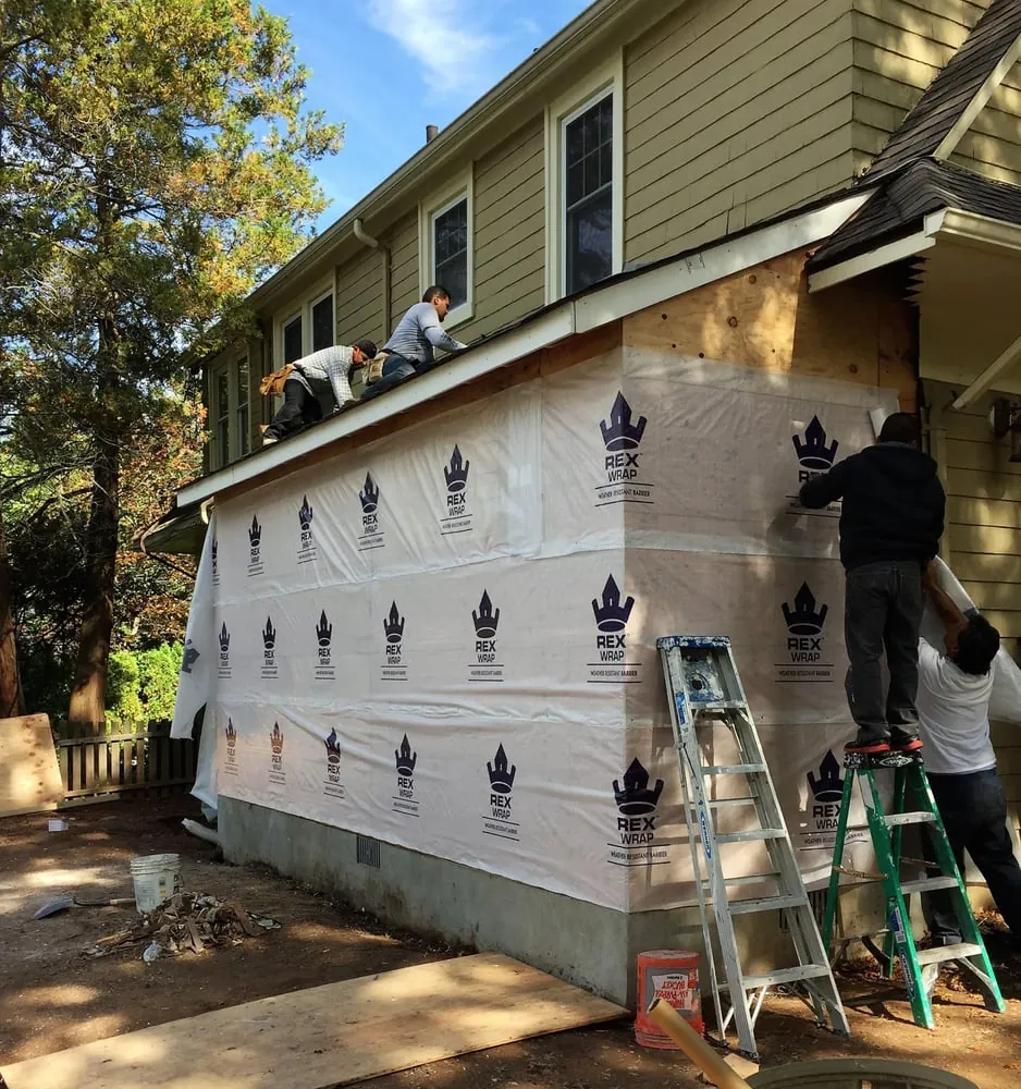 Single-story addition wrapped in white house wrap while a crew works on the roof edge and from ladders.