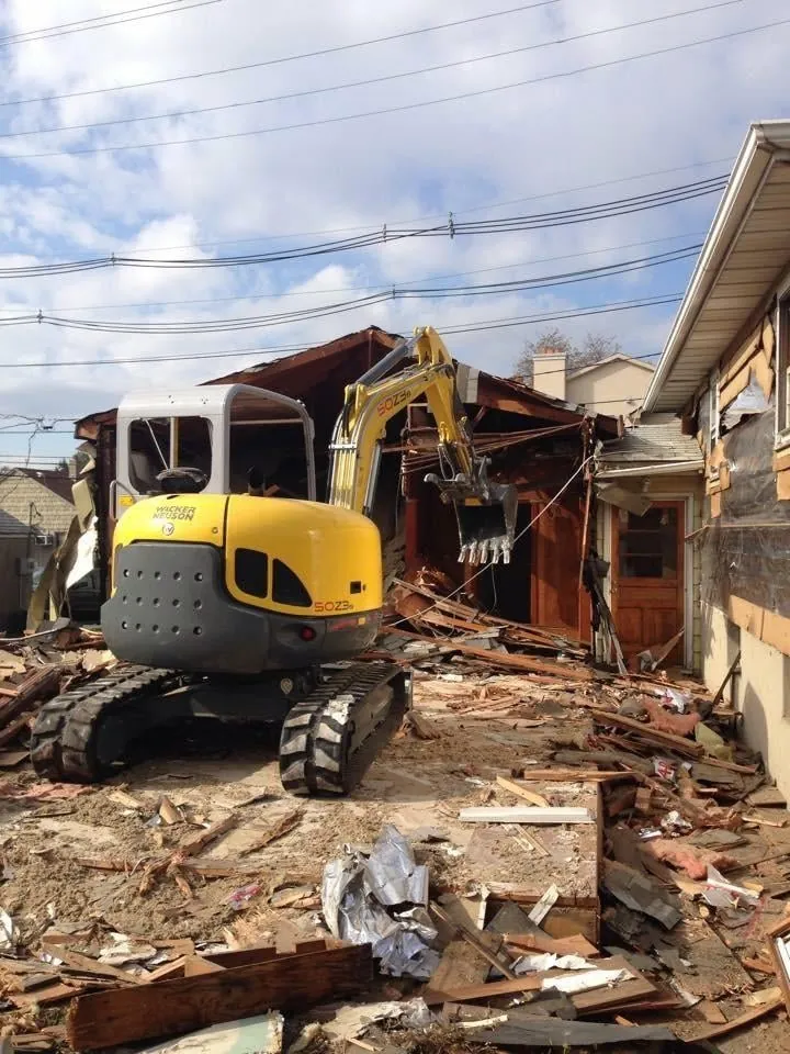 Yellow mini excavator tearing down a wood-framed structure with demolition debris covering the lot.