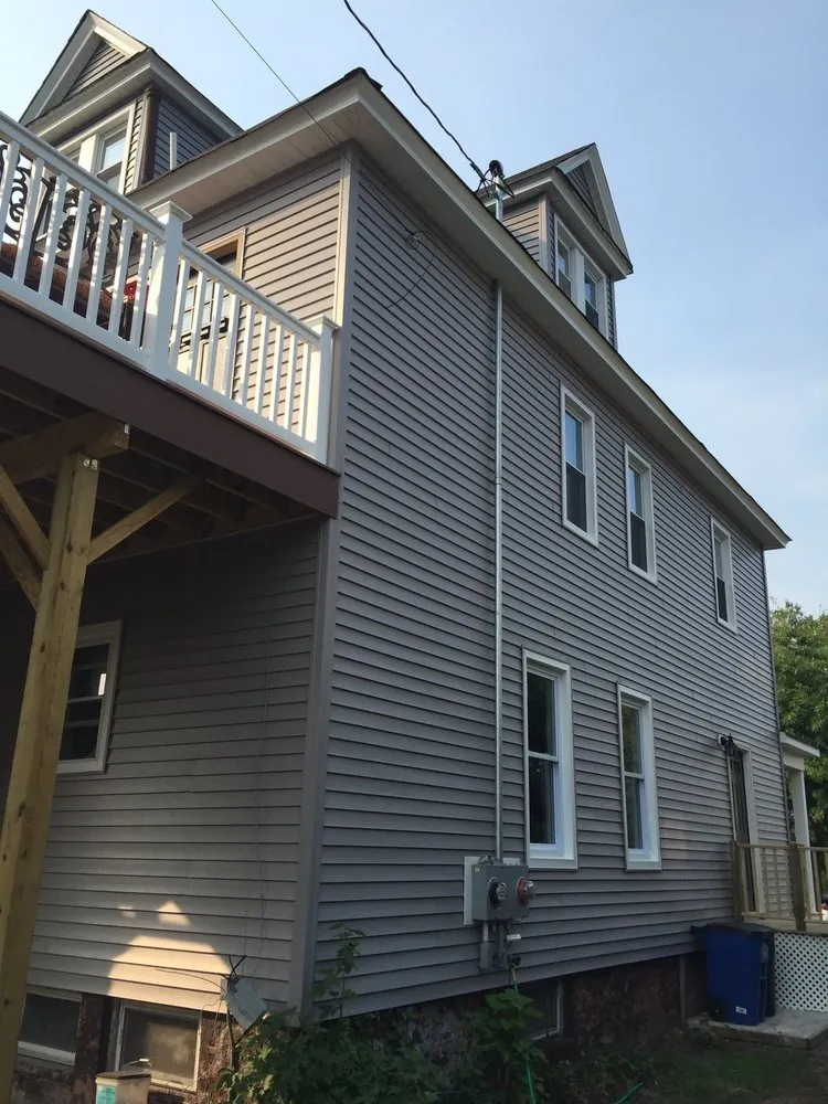 Side wall of a tall house with new gray vinyl siding, white windows and a white-railed deck at the rear corner.
