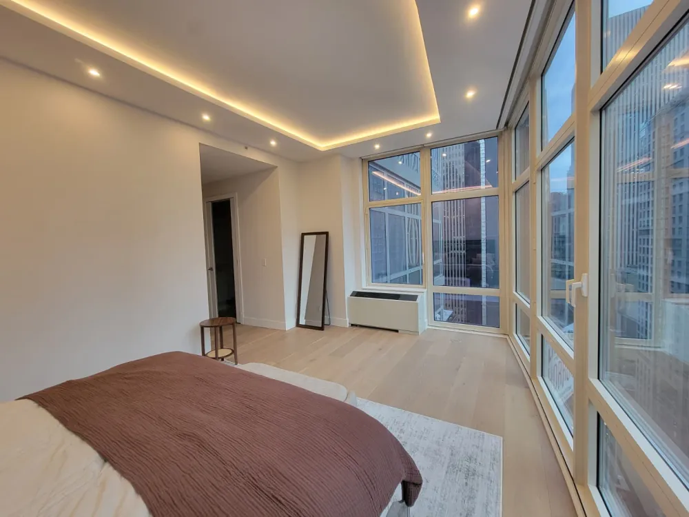 Bedroom with LED cove-lit tray ceiling, recessed lights, light oak floor and a wall of tall windows.
