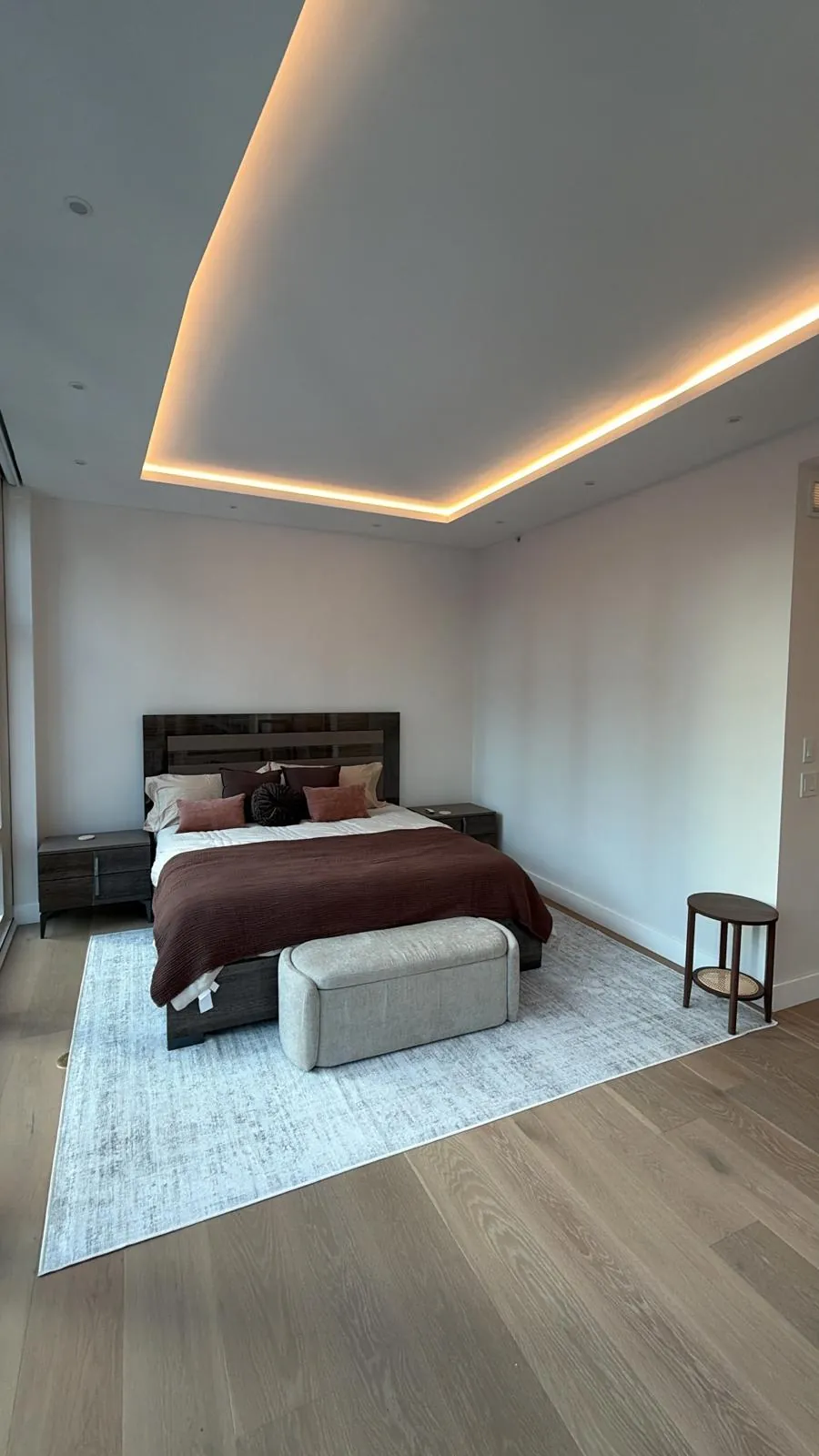Bedroom with recessed tray ceiling lit by warm LED strips, wide plank oak floor, bed and upholstered bench.