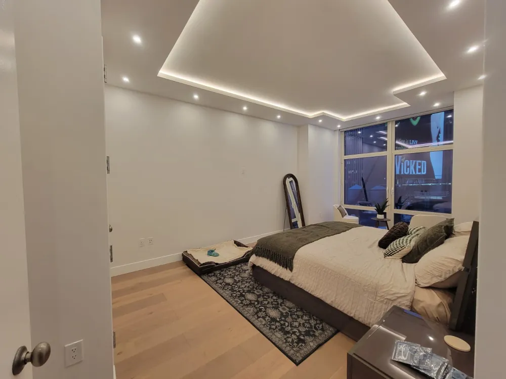 Bedroom with stepped tray ceiling and LED cove lighting, recessed lights, oak floor, patterned rug and city window.