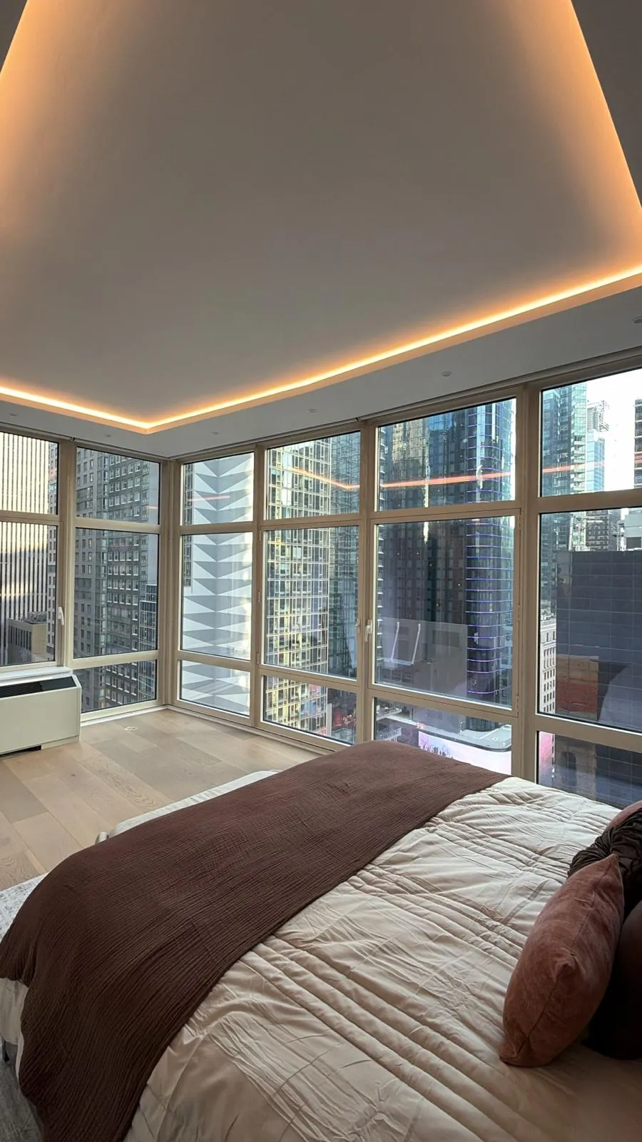 Corner bedroom with tray ceiling and warm LED cove lighting, light oak floor and floor-to-ceiling windows.