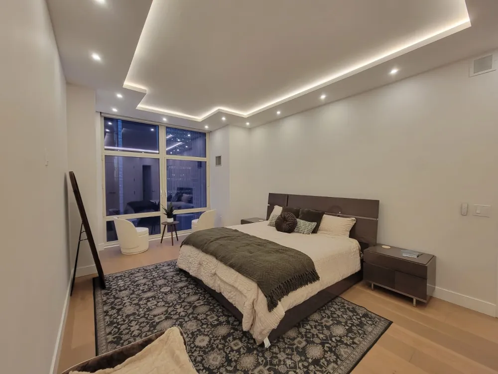 Bedroom with stepped LED tray ceiling, recessed lights, oak floor, patterned rug and a seating area by the window.