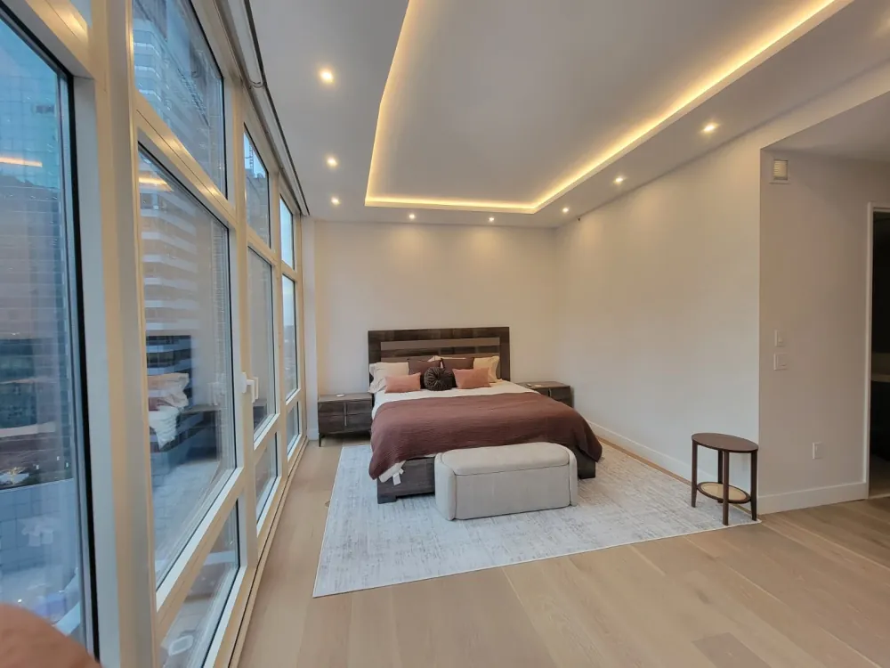Bedroom with LED cove-lit tray ceiling, recessed lights, oak floor and a long wall of windows beside the bed.
