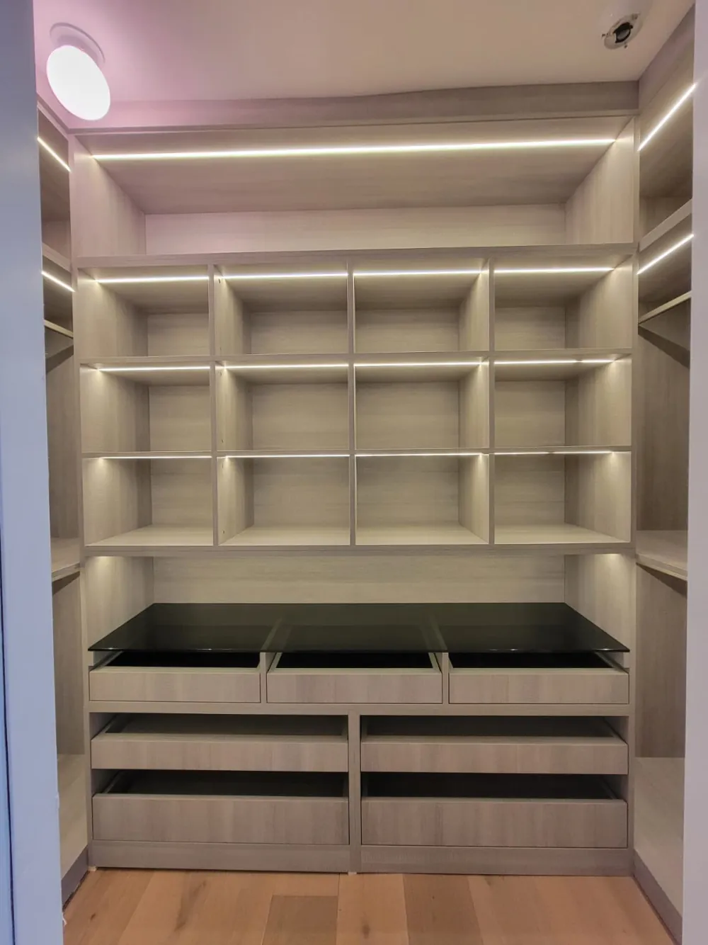 Walk-in closet wall of LED-lit cubbies above built-in drawers with a smoked glass top, hanging sections at the sides.