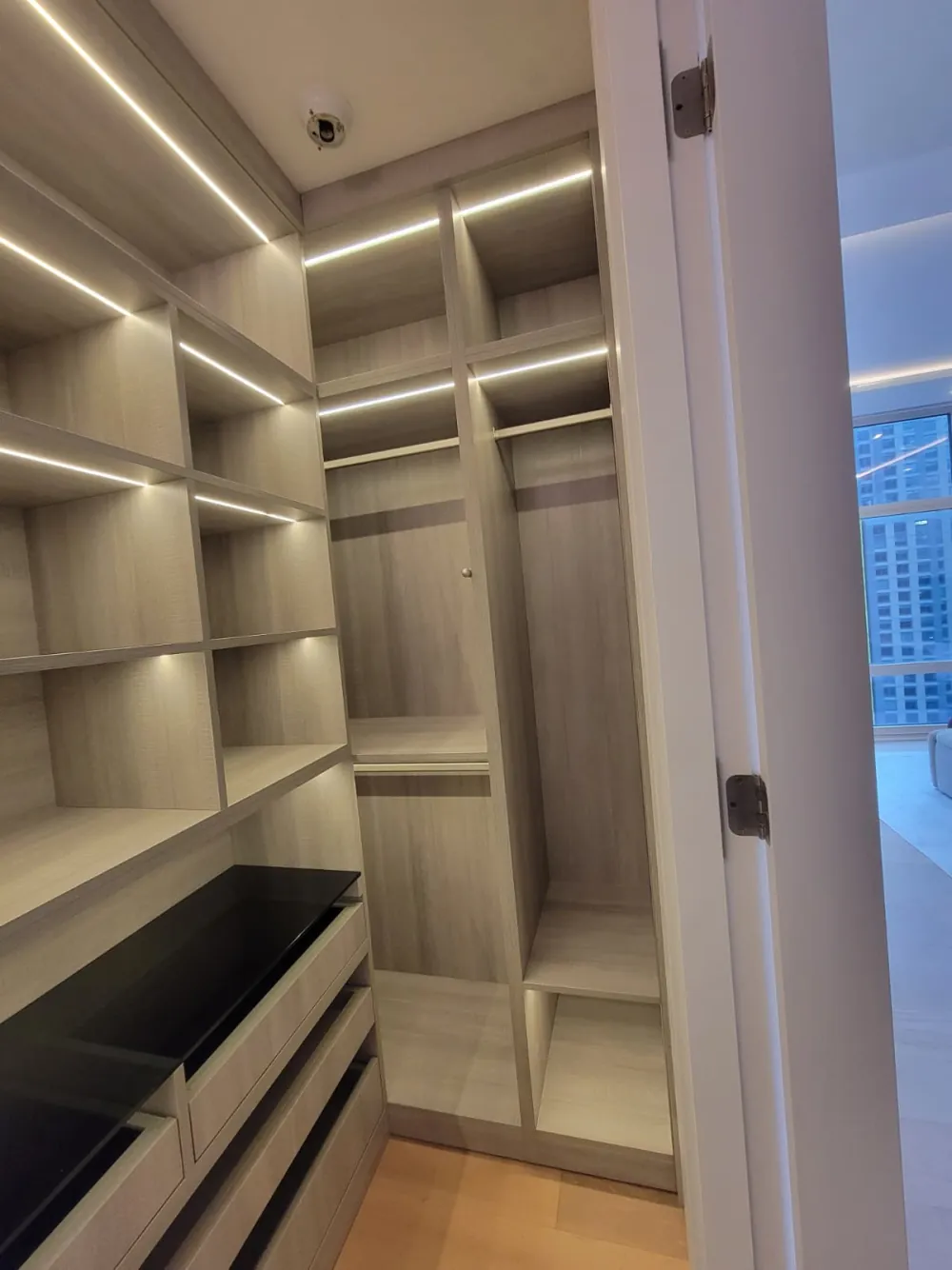 Walk-in closet with wood-grain cubbies, LED shelf lighting, double hanging rods and drawers with a black glass top.