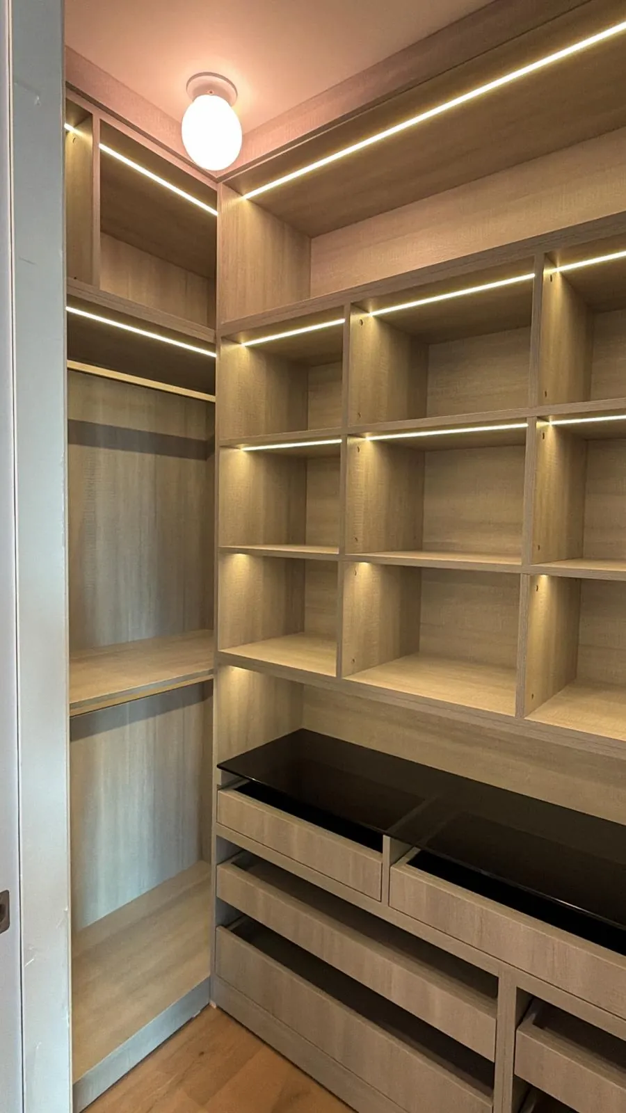 Custom closet with lit cubby shelves, a hanging section and pull-out drawers topped with smoked glass.