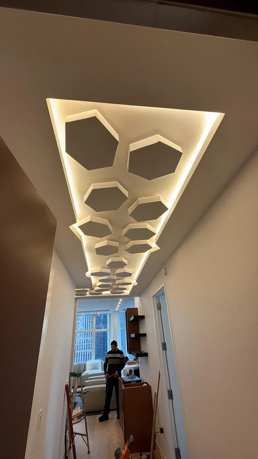 Hallway ceiling with layered hexagon drywall panels and hidden LED lighting, with a ladder and cabinet boxes below.