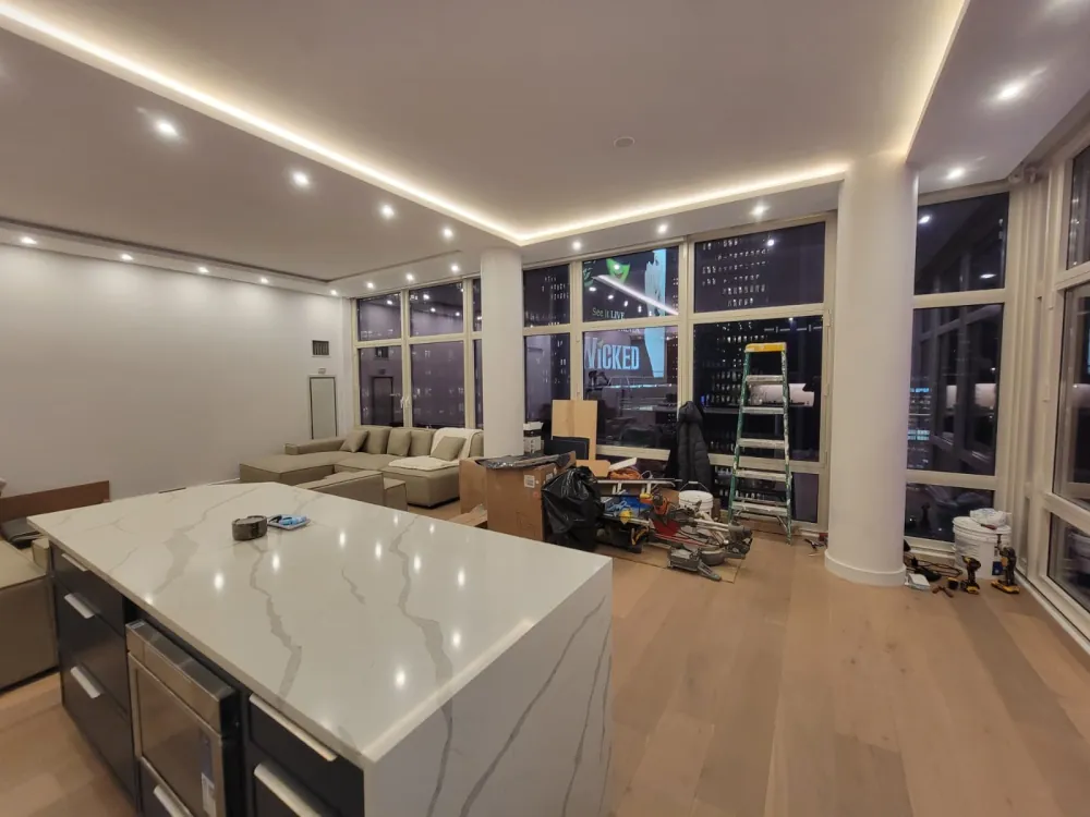 Open living area with LED cove ceiling and round columns, quartz waterfall island in front, tools and ladder by windows.