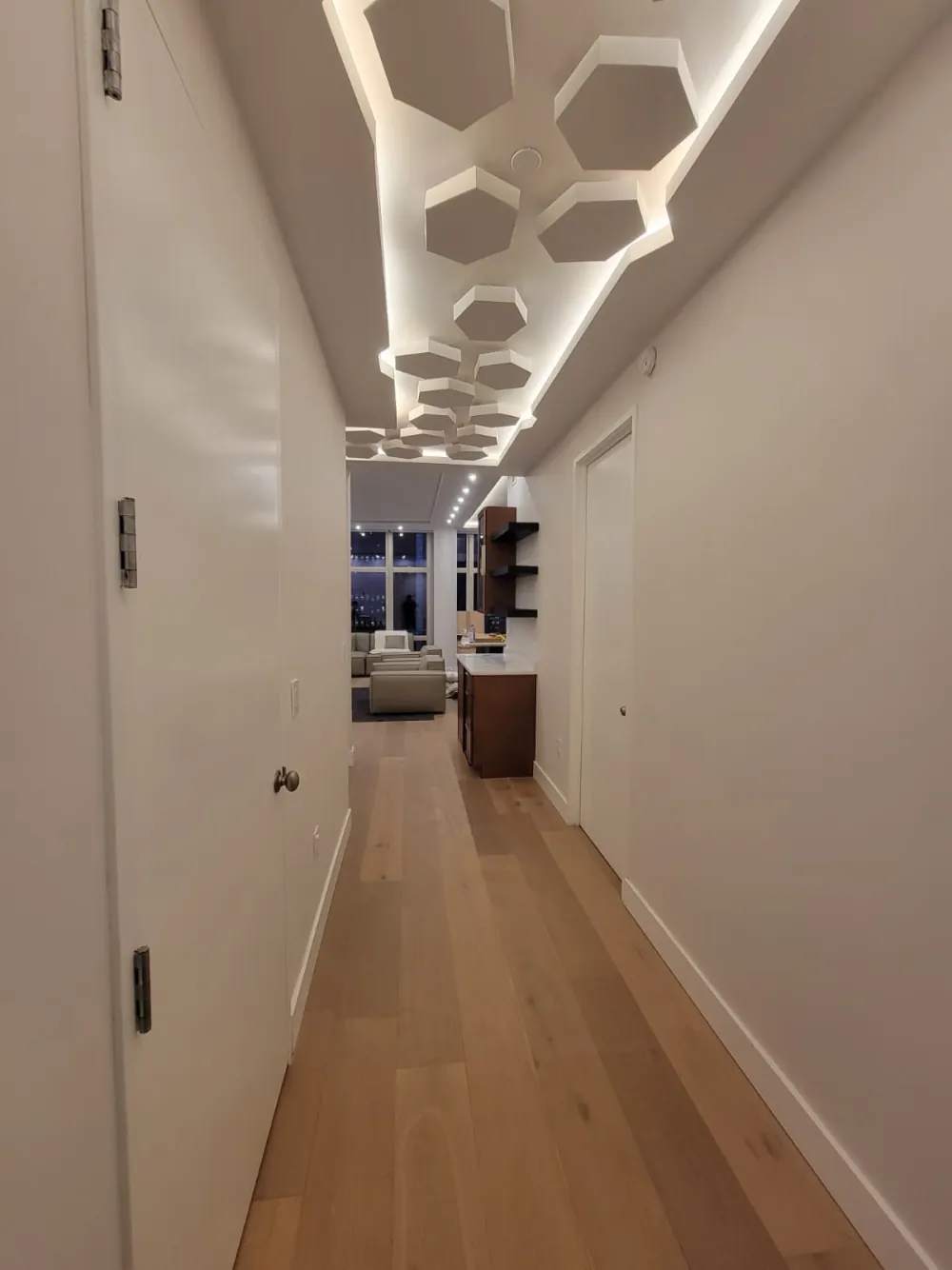 Long hallway with hexagon drywall ceiling panels lit by hidden LED strips, white doors and wide plank oak floor.