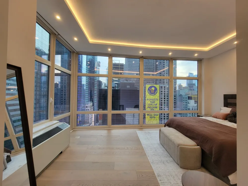 Bedroom with LED cove-lit ceiling, wide plank oak floor and corner floor-to-ceiling windows overlooking city towers.