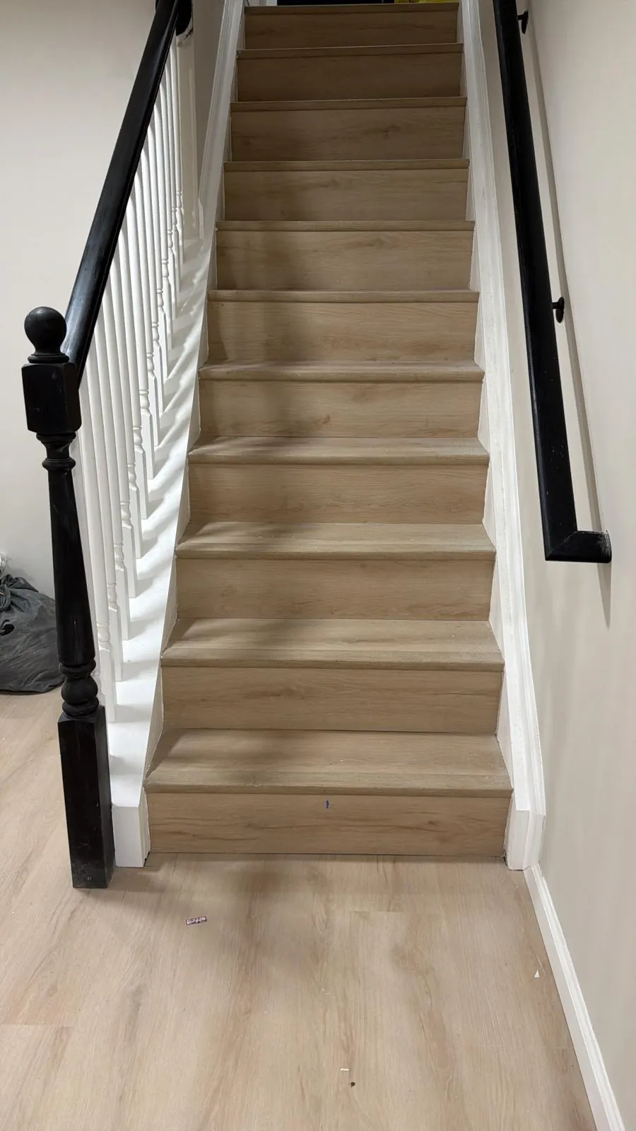 Straight staircase with light oak vinyl treads and risers, white balusters, black newel post and black wall handrail.