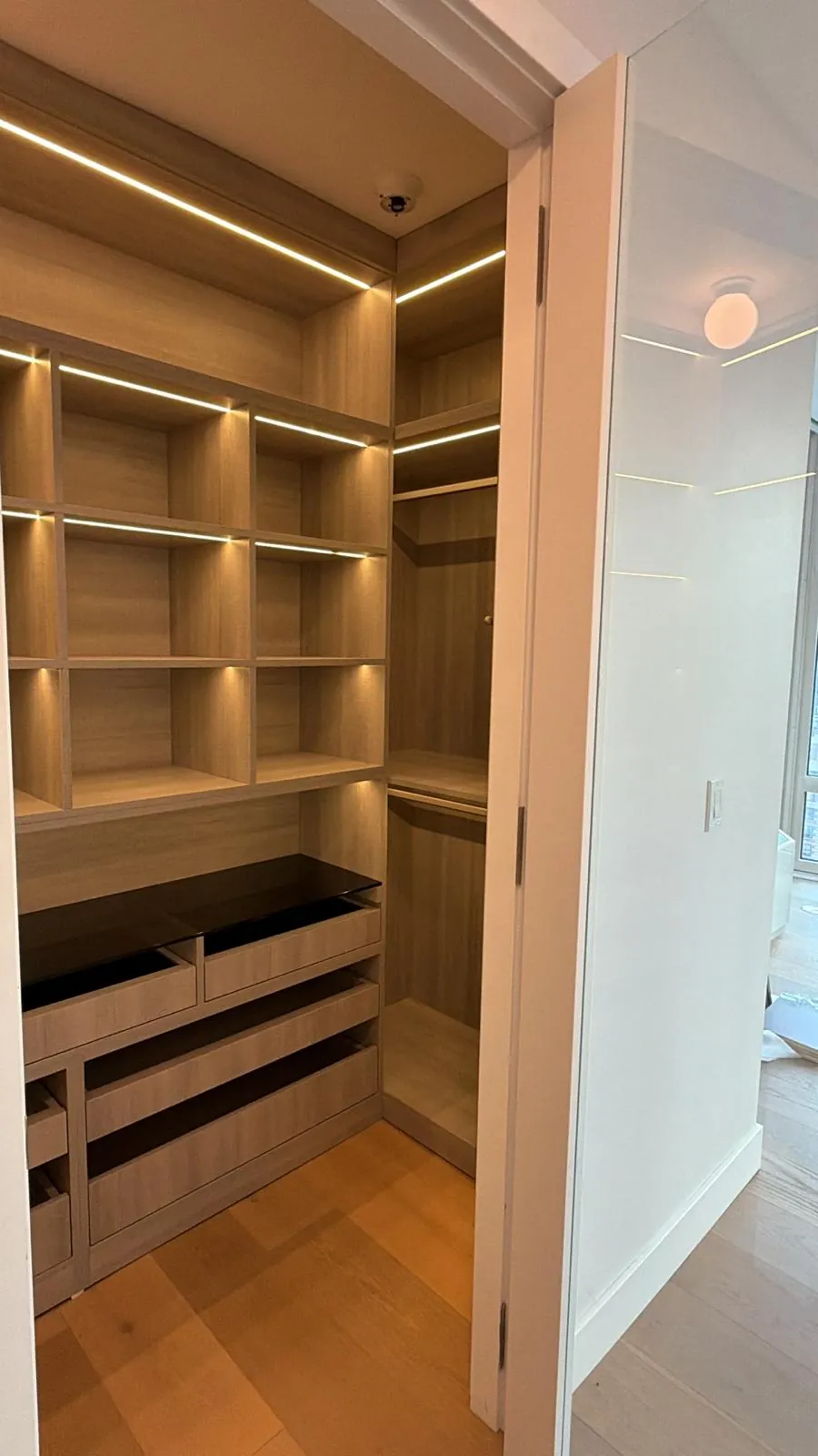 Walk-in closet with light wood-grain shelving, LED strip lighting, hanging rods and drawers under a black glass top.