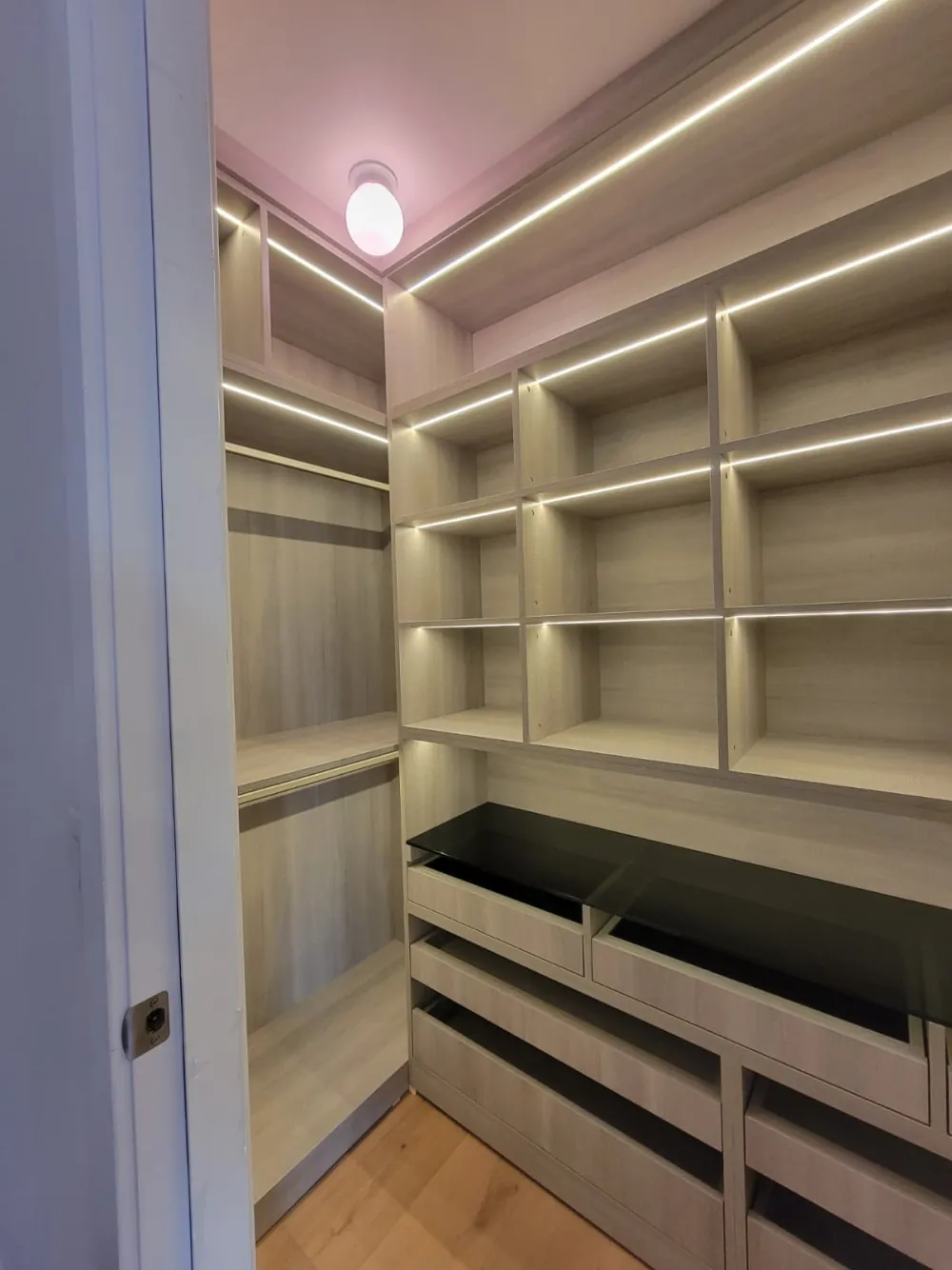 Walk-in closet with LED-lit cubby shelves, a hanging section and wood-grain drawers under smoked glass.