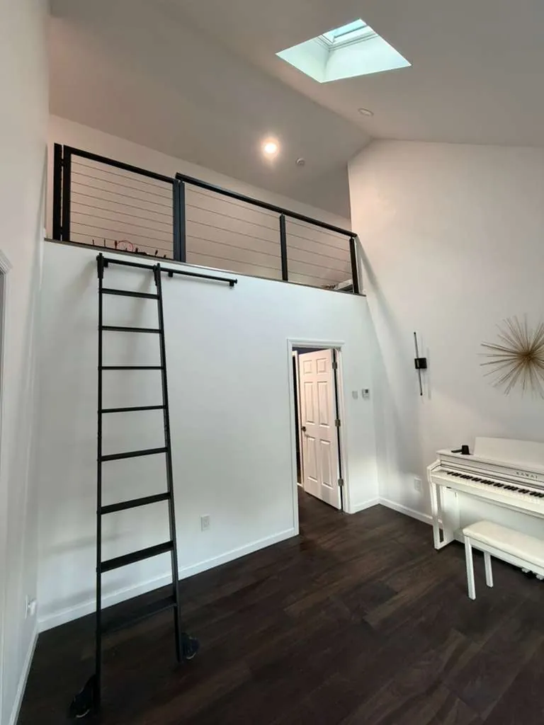 Two-story room with a loft edged by black cable railing, a black rolling library ladder, skylight and dark wood floor.