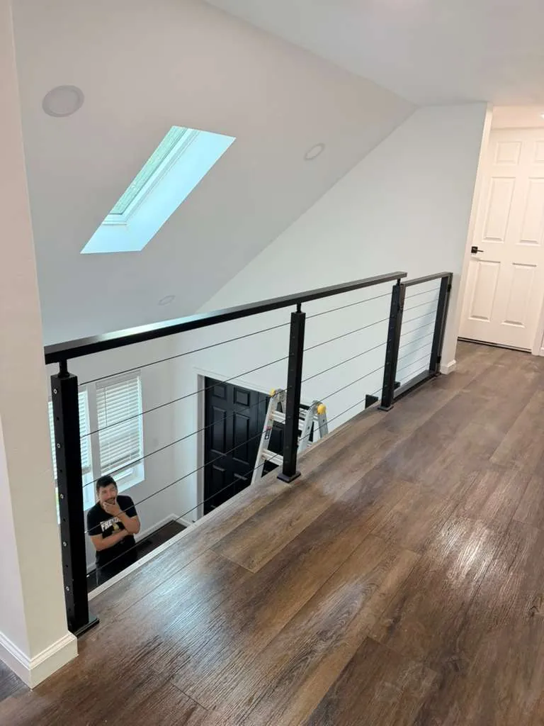 Upstairs loft with wood-look floor, black cable railing overlooking the room below and a skylight in the sloped ceiling.