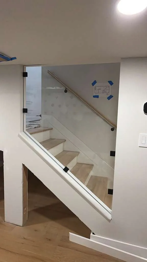 Staircase with white oak treads, a frameless glass guard with black clamps and a wood handrail, with painter's tape still up.